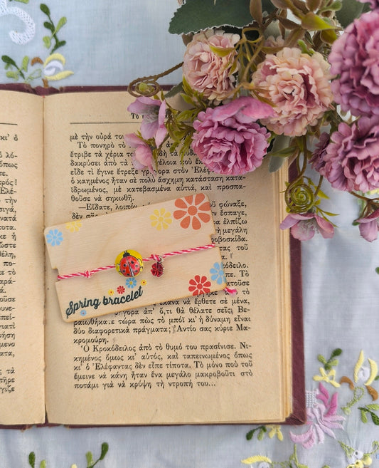 A person's wrist wearing an adjustable bracelet with a ceramic ladybug charm, with red and white string and yellow element, presented in front of an open book and floral background.